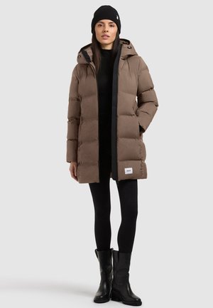 Femme portant une longue doudoune marron avec capuche, leggings noirs, bottes noires et bonnet noir, debout devant un fond uni.