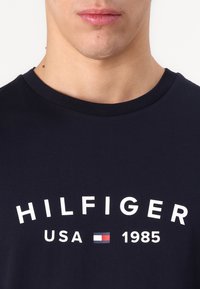 Tamno plava pamučna majica s bijelim zakrivljenim natpisom "HILFIGER USA 1985" i malim grafičkim prikazom trobojnog (crvene, bijele i plave) zastave.