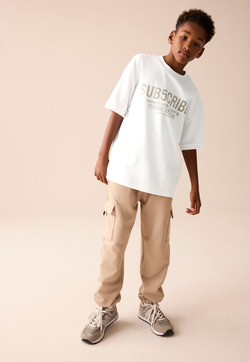Ragazzo con t-shirt grafica bianca oversize, pantaloni cargo beige e sneakers grigie, in piedi davanti a uno sfondo beige uniforme.