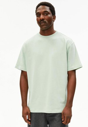 Man met snor die een effen lichtgroen oversized T-shirt met korte mouwen en een donkerbroek draagt, staand tegen een witte achtergrond.