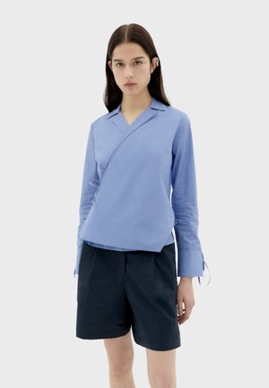 Camisa envolvente azul claro con mangas largas, escote en V y lazos en el costado. Combinada con shorts de corte navy, que ofrecen un ajuste relajado.