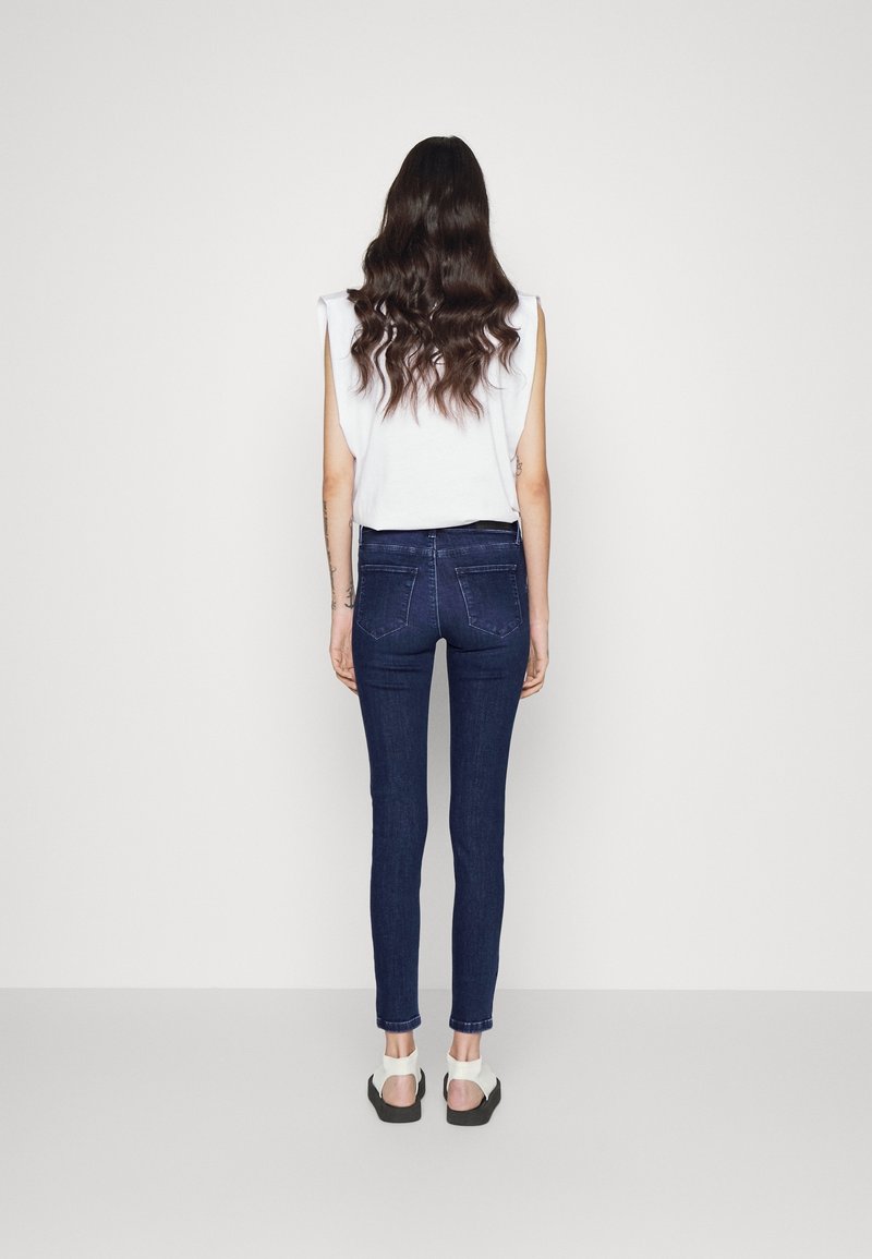 Úzké tmavě modré džíny se zadními kapsami, kombinované s krátkým bílým tílkem bez rukávů a bílými sandály. Hladká textura a přiléhavý střih.