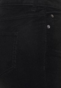 Schwarze Cordhosen mit geripptem Textur, versehen mit Metallknöpfen und aufgenähten Taschen, seitlich betrachtet.