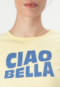 Yellow cotton t-shirt featuring a blue distressed text print saying "CIAO BELLA". Classic crew neck design. Smooth texture.