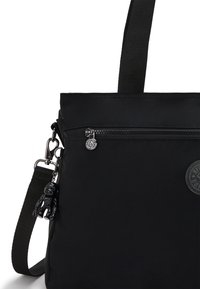 Black nylon tote bag with adjustable strap, zippered pocket, and round logo patch. Features a black bear charm attached to the zipper.