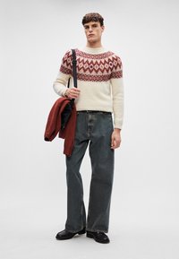 Strickpulli in Creme mit rotem geometrischen Muster, kombiniert mit lockeren, dunklen Denim-Jeans und schwarzen Schuhen, hält eine rote Strickjacke und eine schwarze Tasche.