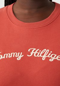 Gros plan sur une personne à la peau foncée portant un t-shirt rouge à col rond avec le texte en cursive blanc "Tommy Hilfiger" sur la poitrine.