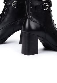 Botas de tobillo de cuero negro con tacón cuadrado, detalles de tachuelas plateadas y cierre de cordones con correas de hebilla. Textura suave en toda la superficie.