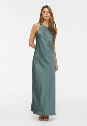 Femeie cu păr lung, purtând o rochie maxi de satin teal fără mâneci și pantofi aurii cu degetele deschise, stând pe un fundal alb.