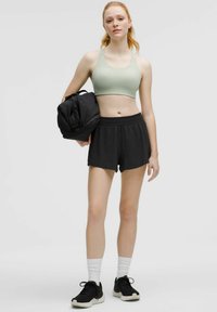 Conjunto deportivo que incluye un sujetador deportivo verde claro y pantalones cortos negros. El modelo sostiene una bolsa de deporte negra. Los calcetines son blancos y los zapatos son negros.