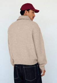 Maglione beige lavorato a maglia con collo alto, trama a coste e vestibilità corta, abbinato a jeans di denim scuro e un berretto color bordeaux.