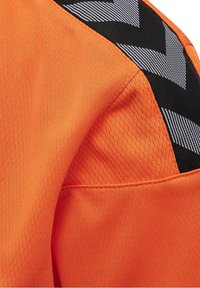Jersey atlético naranja con tejido texturizado, que presenta acentos de franjas en forma de chevron en blanco y negro en el hombro y detalles de costura contrastantes.
