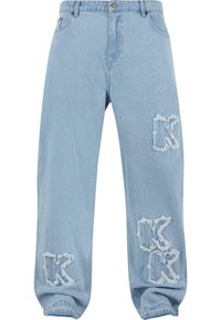 Lyseblå, slitt denimjeans med løs passform, som har frynsete kanter og to fremtredende "K"-lapper på fronten.