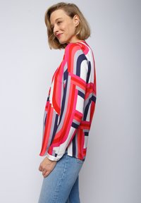 Bunte Bluse mit auffälligen geometrischen Mustern in Rot, Pink und Marine, langen Ärmeln und leichtem Stoff, kombiniert mit hellblauen Jeans.