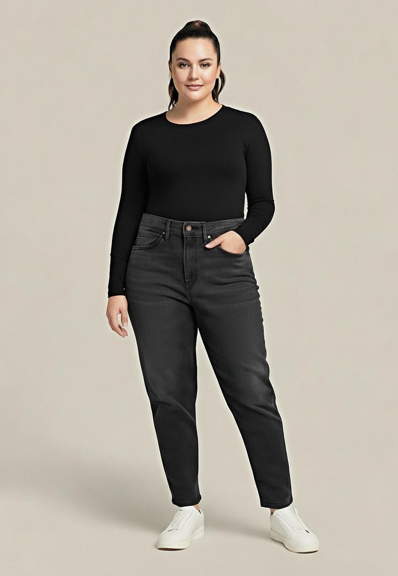 Kobieta stoi z jedną ręką w kieszeni, ubrana w czarną koszulę z długim rękawem, ciemne dżinsy i białe trampki na beżowym tle.