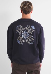 Sweatshirt marine avec motif floral brodé en bleu clair et beige, centré au dos. Manches longues, col rond, coupe standard.