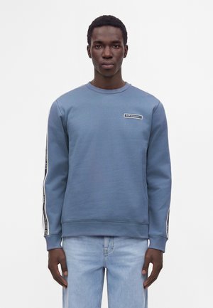 Jeune homme portant un sweat-shirt bleu avec un ruban de marque sur les manches et un petit patch logo sur la poitrine, associé à un jean bleu clair, debout immobile.