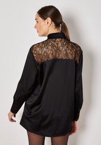 Blusa nera con schiena in pizzo trasparente, maniche lunghe con polsini a bottone e un frontale in satin liscio per un contrasto di texture.