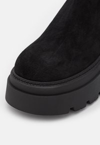 Black suede ankle boot with a chunky rubber sole, round toe, and visible stitching detail around the sole. Textured surface.