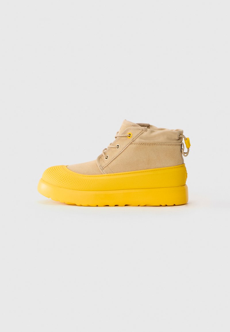 Bottines montantes beige avec semelle et capuchon en caoutchouc jaune vif, devant à lacets et col réglable à cordon, sur un fond blanc.