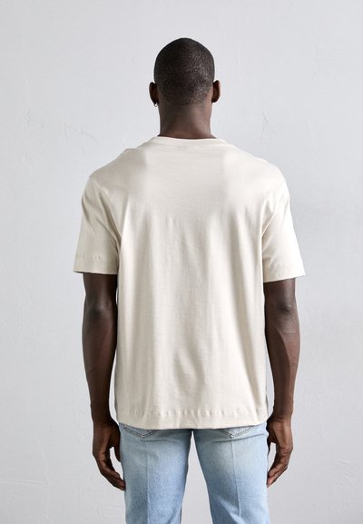 μπεζ βαμβακερό μπλουζάκι σε χαλαρή εφαρμογή, κοντά μανίκια και στρογγυλή λαιμόκοψη, συνδυασμένο με ανοιχτό μπλε τζιν παντελόνι.