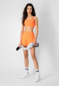Sujetador deportivo naranja neón y pantalones cortos a juego, hechos de un tejido elástico, con una cintura elástica y cordón, combinados con zapatillas blancas y calcetines.