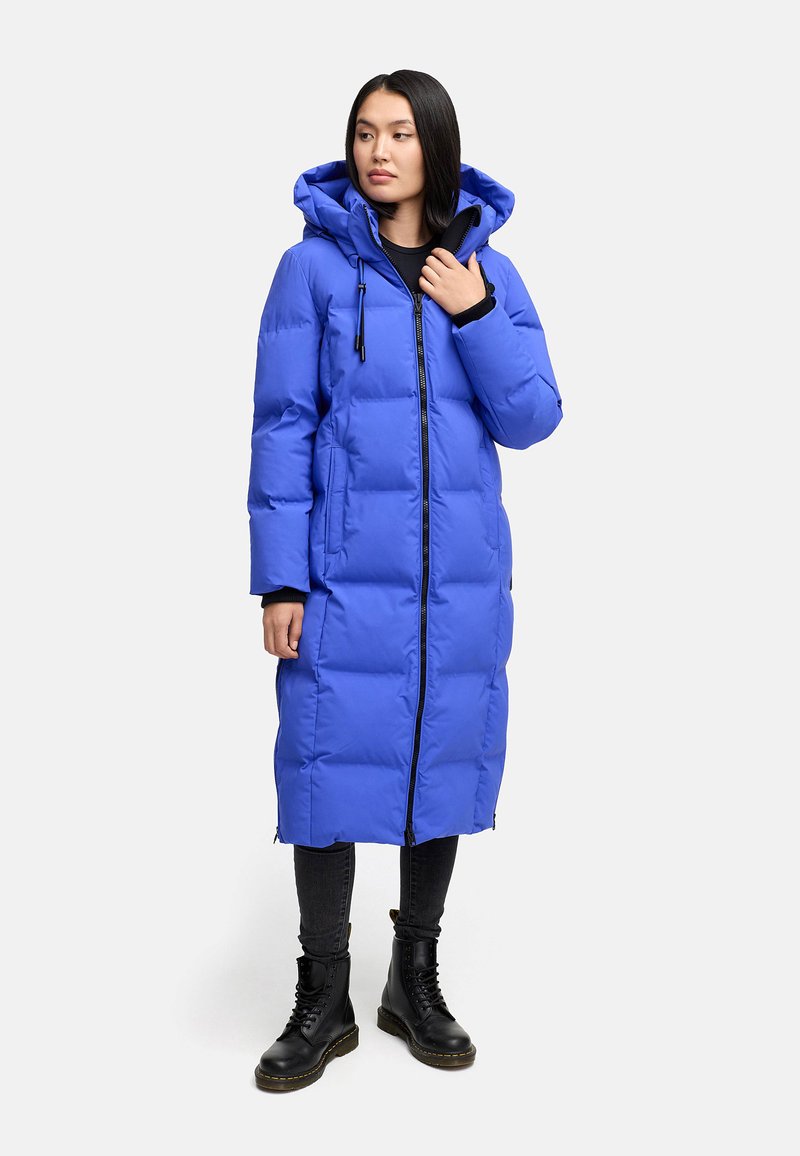 Lang pufferjas in een levendige blauwe kleur, met een capuchon, ritssluiting en een gestikte textuur. Draagt met zwarte jeans en veterschoenen.
