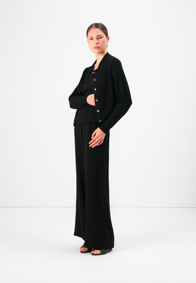 Woman standing in black buttoned cardigan, black top, wide-leg black pants, and black open-toe shoes against a plain white background.