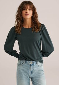 Sötétzöld, hosszú ujjú ing puffos ujjal, kerek nyakkivágással és puha anyagból, világoskék, magas derekú farmerral párosítva.