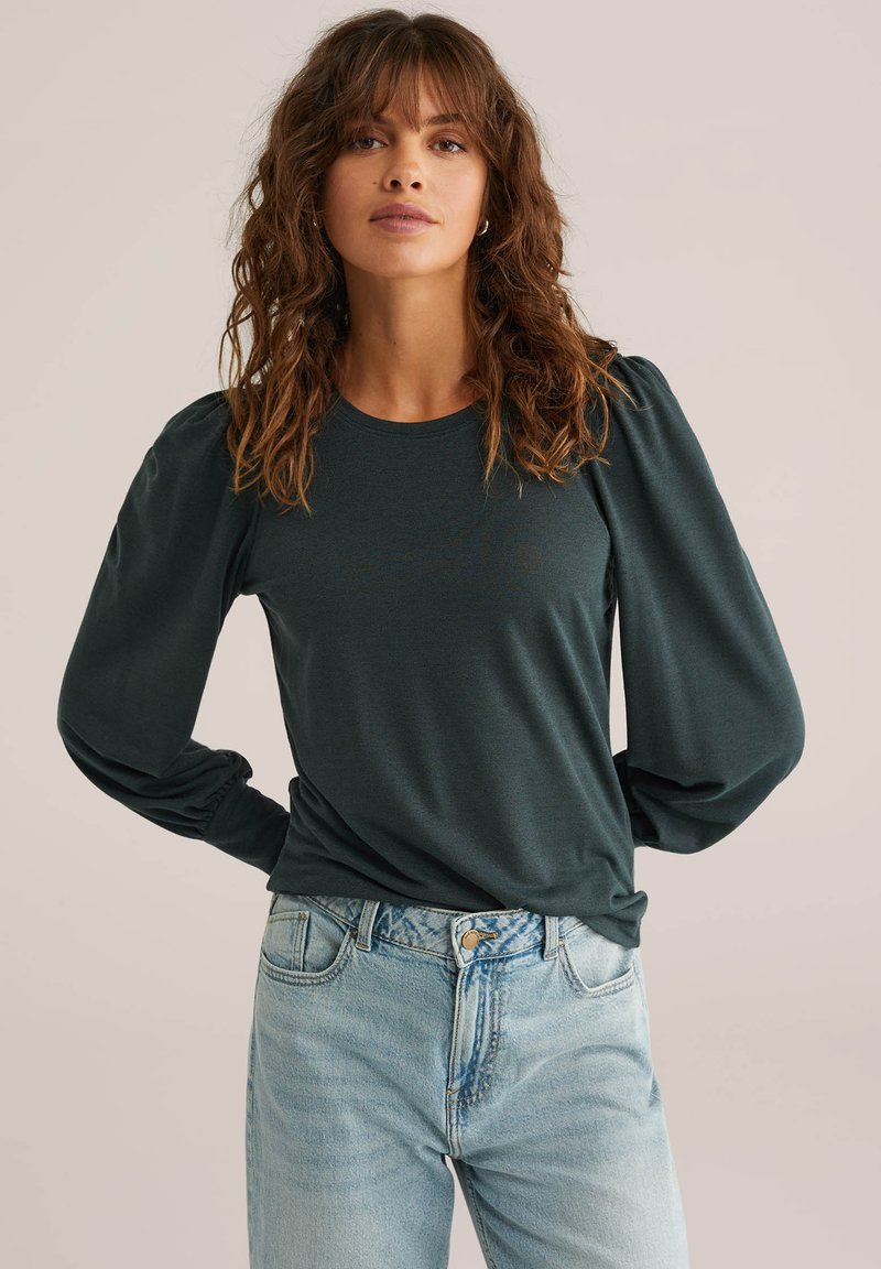 Sötétzöld, hosszú ujjú ing puffos ujjal, kerek nyakkivágással és puha anyagból, világoskék, magas derekú farmerral párosítva.