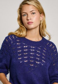 Lila stickad tröja med ett mönstrat ok och öppna vävdetaljer, rund halsringning och uppvikt ärm. Mjuk textur och lös passform.