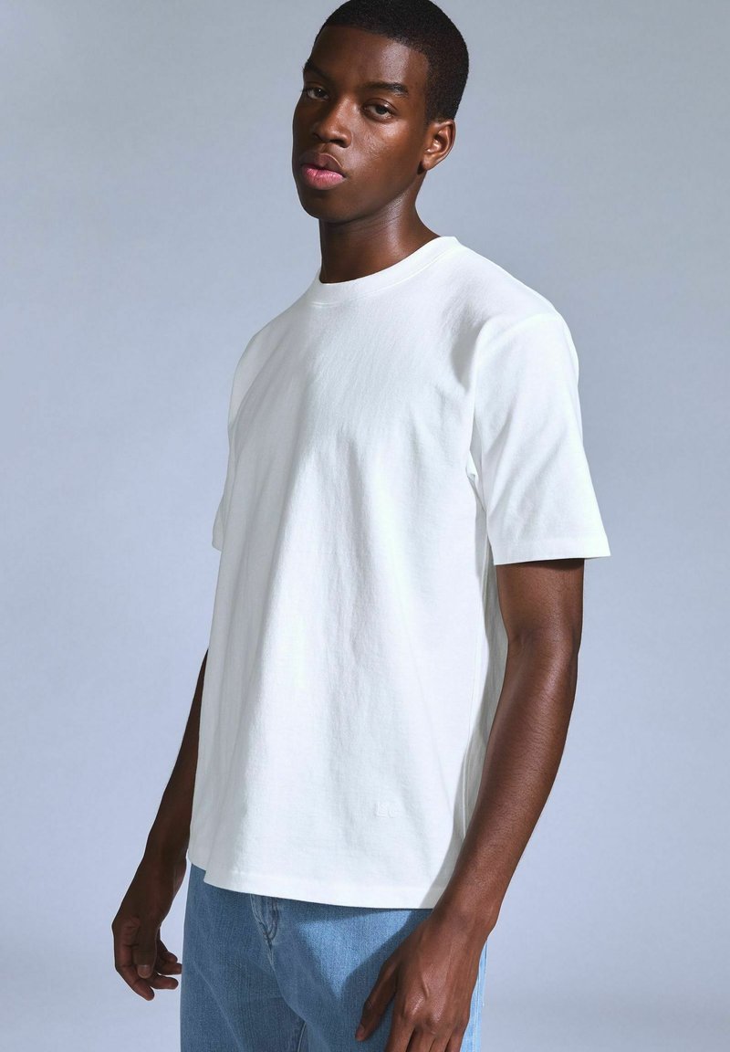 White cotton T-shirt with a round neckline and short sleeves. Loose fit with minimal detailing. Model poses against a gray background.
