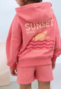 Różowy komplet z bluzy z kapturem i szortów z napisem "SUNSET BEACH" oraz grafiką słońca i fal; miękka faktura, luźny krój, ściągacze przy mankietach.