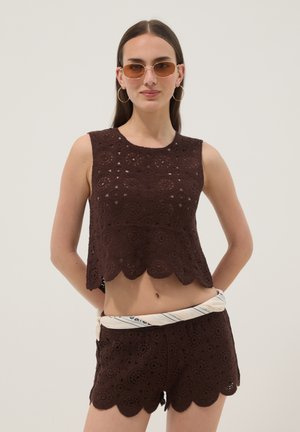 Mujer con top sin mangas de crochet marrón y pantalones cortos a juego con bordes festoneados, gafas de sol, pendientes de aro y un cinturón ligero de pañuelo.