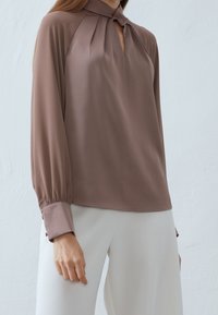 Braune langärmelige Bluse mit hohem Kragen und plissiertem Detail aus glattem Stoff, getragen zu weißen Hosen, mit einem entspannten Schnitt.