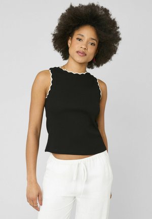 Femme aux cheveux bouclés portant un haut noir sans manches avec bordure blanche et un pantalon blanc à cordon, debout devant un fond gris.