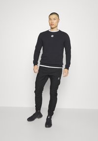 Felpa nera con scollo rotondo e logo sul petto, abbinata a pantaloni sportivi neri a gamba affusolata con un piccolo logo. Indossata con scarpe nere.