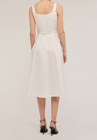 Witte katoenen jurk met een aansluitend lijfje, A-lijn rok en een verstelbare riem. Heeft een ritssluiting op de rug en schouderbandjes. Groene hakken getoond.