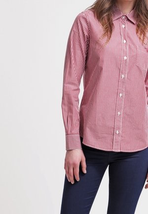 Femme portant une chemise à rayures verticales rouges et blanches boutonnée, à manches longues, et un pantalon bleu foncé, debout devant un fond uni.