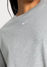 Světle šedé tričko vyrobené z měkké bavlny, s kulatým výstřihem a malým bílým logem Nike na hrudi. Hladká textura a jednoduchý design.