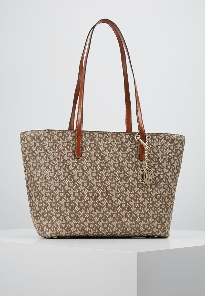 Sac fourre-tout en toile beige avec un motif géométrique marron. Doté de anses en cuir marron et d'un charm en métal doré accroché.