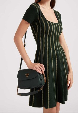 Femme portant une robe vert foncé à rayures verticales, tenant un sac à épaule noir Guess avec des détails en chaîne dorée.