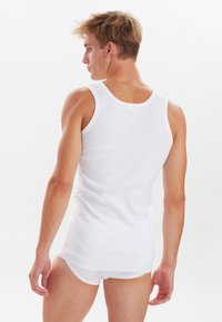 Hvid bomulds tanktop med rund hals og smalle stropper, tætsiddende design, minimal tekstur, båret over hvide trusser.