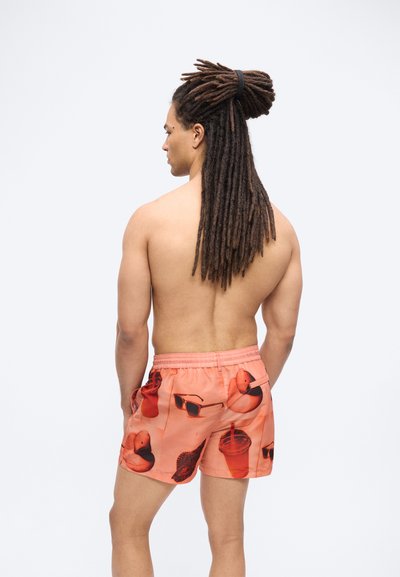 Homme aux longues dreadlocks attachées, torse nu, portant un short de bain corail imprimé de motifs de lunettes de soleil, bouées en forme de canard, feuilles et gobelets, vu de dos.