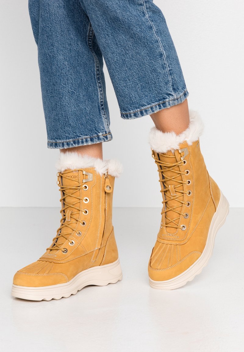 Bottes d'hiver marron clair à lacets avec bordure en fourrure blanche portées avec un jean court bleu sur un sol blanc.