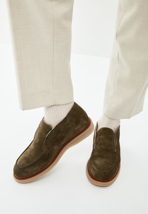 Personne portant un pantalon beige et des chaussettes blanches avec des chaussures en cuir suédé vert foncé à enfiler, semelles marron clair, sur un sol blanc.