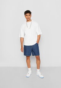 Tricou alb cu mânecă scurtă, pantaloni scurți din denim albastru, adidași albi cu accente albastre, stând pe un fundal gri. Colier din lanț vizibil.