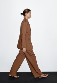 Brązowy lniany garnitur z podwójnym zapięciem w marynarce i spodniami o szerokich nogawkach. Noszony z brązowymi sandałami. Gładkie tło w stonowanych kolorach.