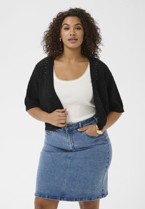 Göndör hajú nő fekete kötött kardigánban, fehér trikóban, kék farmer szoknyában, arany karika fülbevalóval és arany karkötővel.