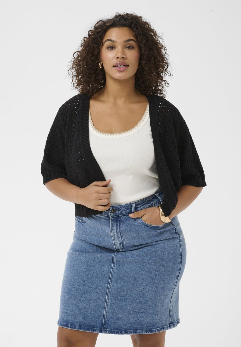 Femme aux cheveux bouclés portant un cardigan en maille noir, un débardeur blanc, une jupe en denim bleu, des boucles d'oreilles créoles en or et un bracelet en or.